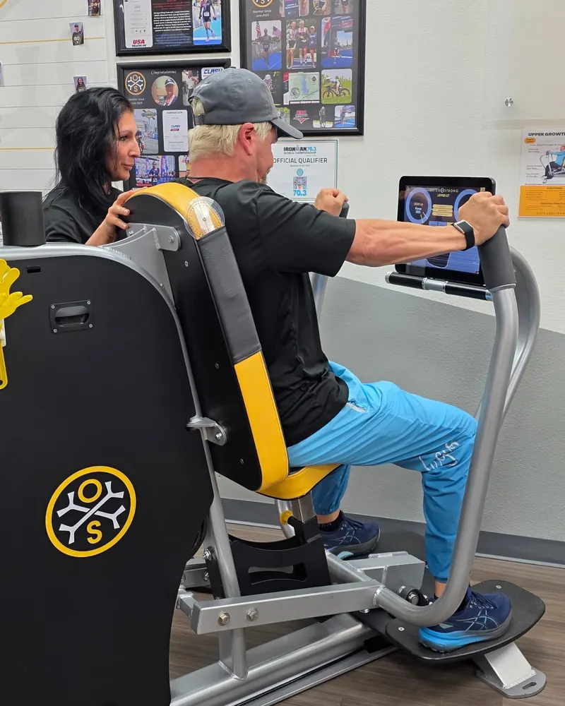 A coach steadies the seat back while a member presses forward on the upper body station, his force on the screen